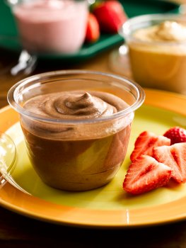 Food photograph of a portion of fluffy chocolate flavour Angel Delight, served in a transparent plastic container with sliced strawberries; served on an orange and yellow plate in a care home setting, with pots of strawberry and butterscotch flavour Angel Delight in the background