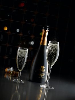 Drinks photograph of two sparkling glasses alongside an open bottle of prosecco, shot on a reflective black background in a dark set with a bottle rack in the background